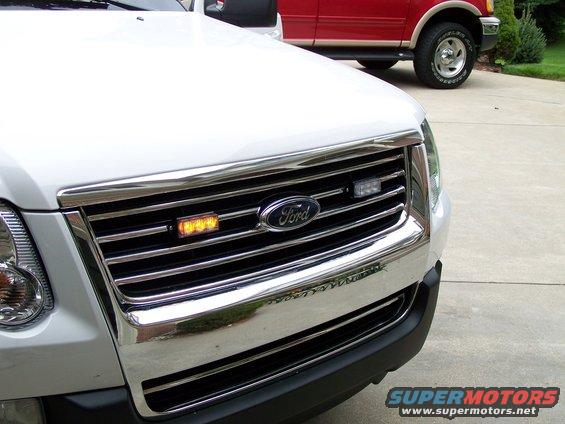 Front grille of a white Ford Explorer with emergency lights installed. A red Ford pickup truck is parked behind it