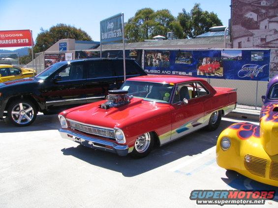 Red hot rod coupe with a large engine blower on the hood, parked next to a black SUV and a yellow custom car
