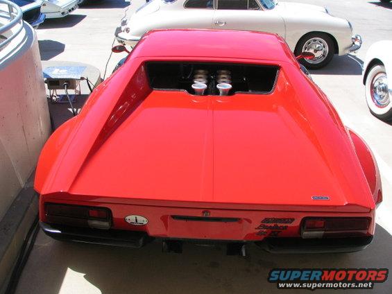 Red sports car with twin exhaust pipes visible in the engine bay. Other cars are parked nearby