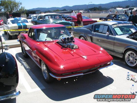Red hot rod with a large blower on the hood, parked in a lot with other cars