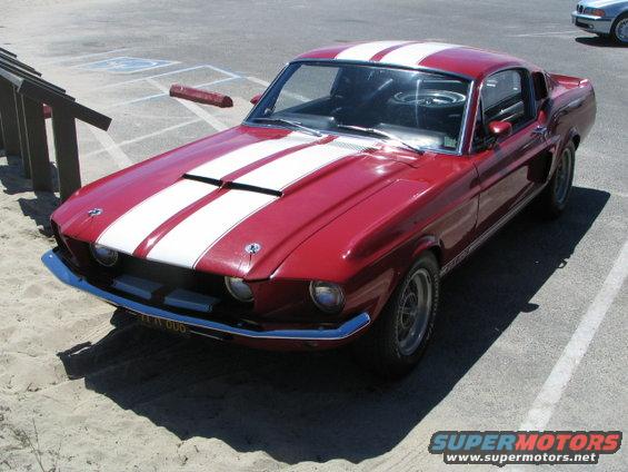 Red 1967 Ford Mustang Shelby GT500 with white racing stripes parked on sand, front 3/4 view