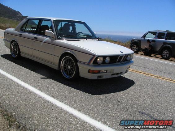 White BMW sedan with red and blue pinstripes, driving on a coastal road