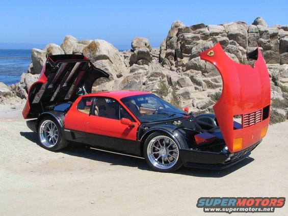 Red and black Ferrari Testarossa with hoods open, parked on a dirt road near rocky coastline