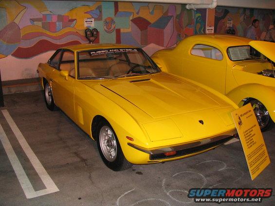 Yellow Lamborghini Urraco parked next to a yellow hot rod. Sign in front of Urraco