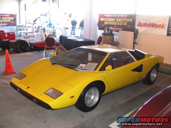 Yellow Lamborghini Countach parked indoors, with a race car and auction banners in the background