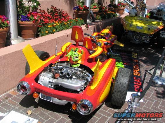 Custom red and yellow hot rod with exposed V8 engine and cartoon mouse mascot. Another yellow vehicle is visible behind it