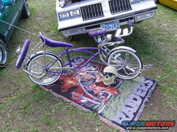 Purple lowrider bicycle with chrome accents, skull with sunglasses, and Raiders blanket. Silver car in background