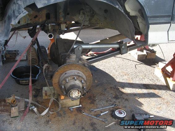 Truck's front suspension disassembled with tools on the ground. Parts are supported by blocks and straps