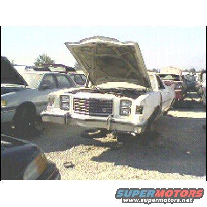 White car with hood open in a junkyard, front view. Grille and headlights visible