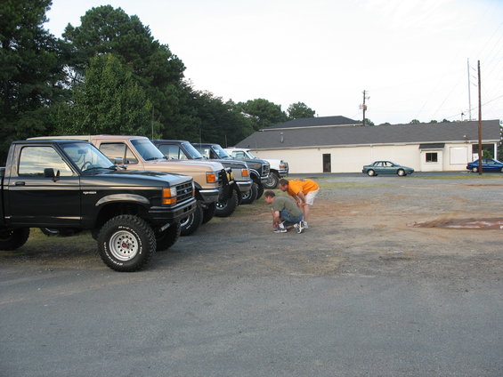 Several pickup trucks parked in a lot, with two guys looking at something on the ground