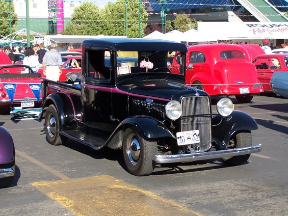 Black 1932 Ford pickup truck with pink pinstripes, parked at an outdoor car show. Red car behind it