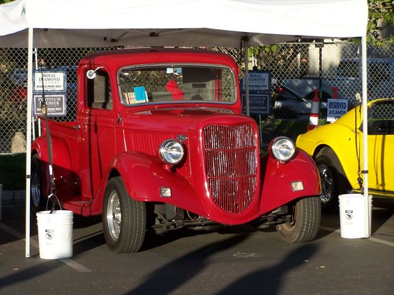 Red vintage pickup truck front view under a tent, with a yellow car to the right