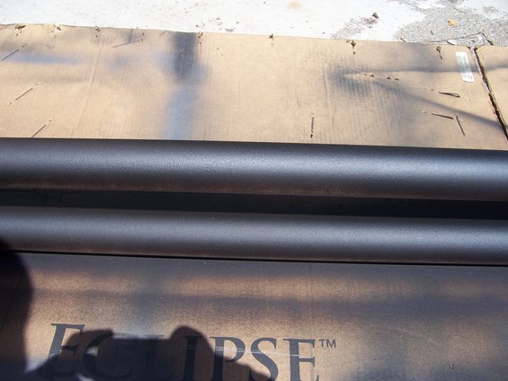 Two dark metal tubes, possibly exhaust pipes or side steps, resting on cardboard with spray paint overspray. Text 'Eclipse' is partially visible