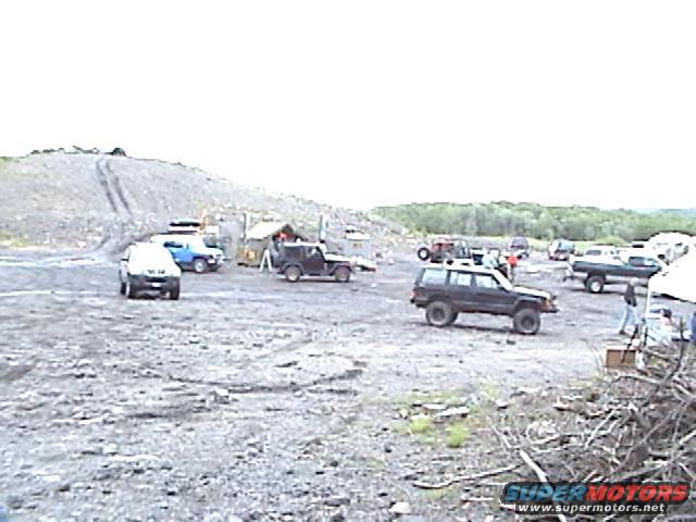 Several off-road vehicles are parked in a gravel lot with tents and trees in the background