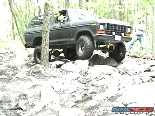 Dark gray 1978 Ford Bronco driving over rocks in a wooded area