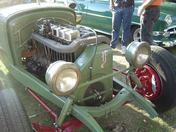 Green vintage car front end with exposed engine and dual headlights. Another green car is visible behind it