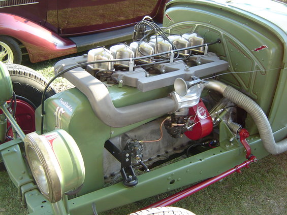 Close-up of a green Franklin engine with a red component and chrome horn. Another car is visible in the background