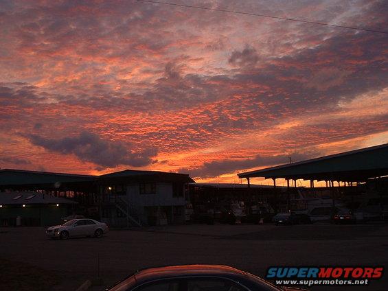 Sunset sky with orange and pink clouds over buildings and boats docked nearby. A few cars are visible in the foreground and background