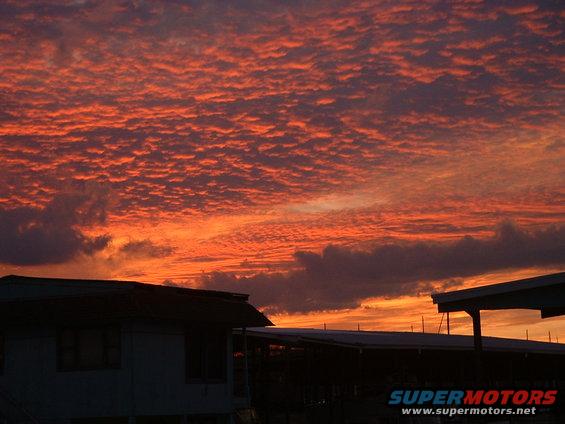 Sunset sky with orange and red clouds over silhouetted buildings and a Supermotors.net watermark