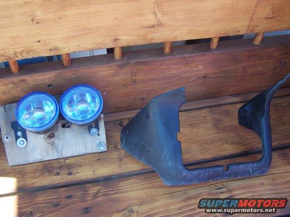 Two blue fog lights mounted on wood, next to a dark car bumper piece