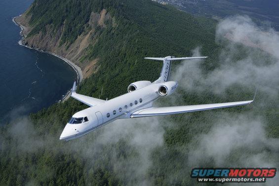 White private jet flying over a forested coastline, with clouds obscuring the land below
