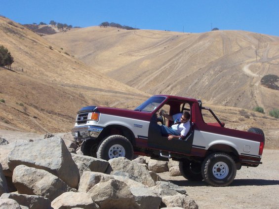 Maroon and white 1991 Ford Bronco off-roading over rocks, driver's door removed