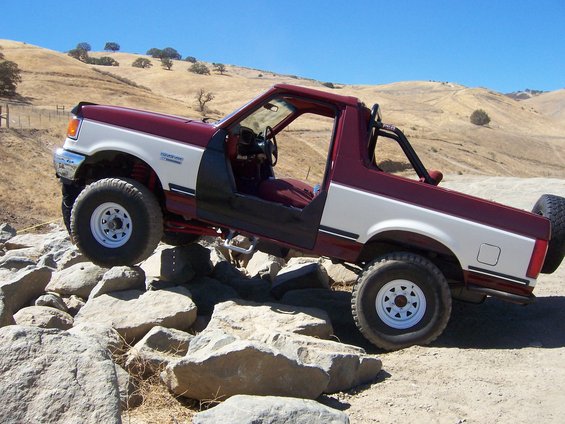 Maroon and white 1991 Ford Bronco off-roading over rocks, driver's door open