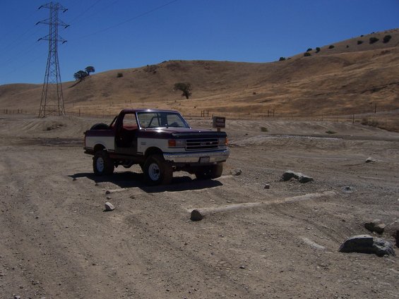 Maroon and white Ford Bronco parked on a dirt road with hills and a power line tower in the background