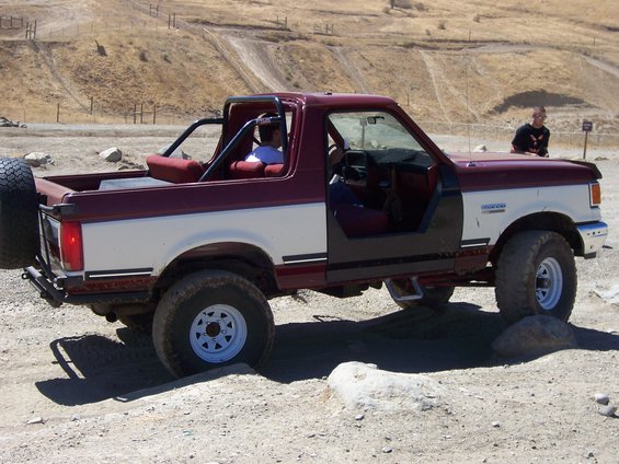 Maroon and white Ford Bronco with doors removed, driving over rocks on a dirt trail. Spare tire mounted on the back