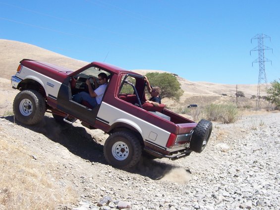 Red and white off-road SUV with lifted suspension and large tires driving on a dirt hill. Two people are inside