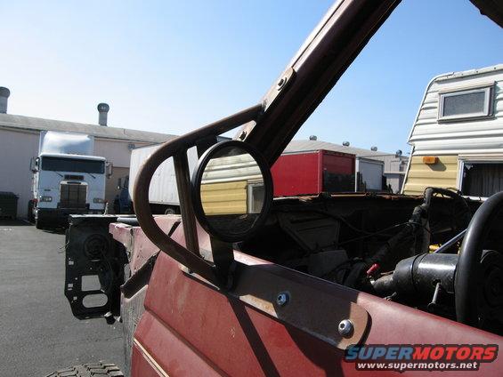 Interior of a red off-road vehicle with a roll cage and side mirror. A semi-truck and trailers are in the background