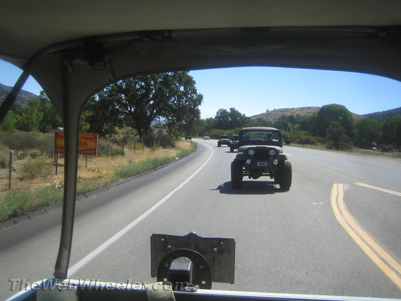 Front view of a dark green Jeep CJ-7 driving on a highway, with other Jeeps behind it