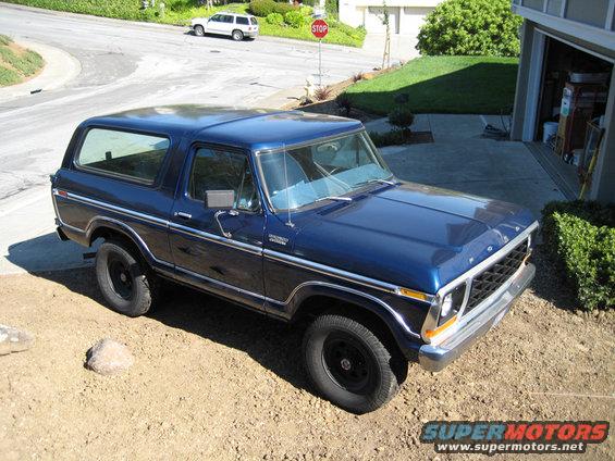1978 Ford Bronco parked in a driveway, blue paint, chrome trim, black wheels. White SUV visible on street