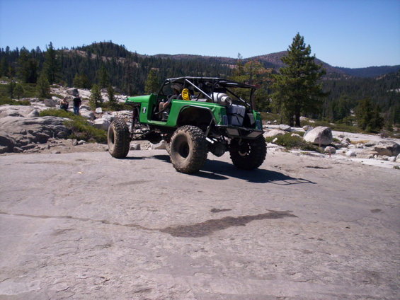 Green off-road buggy with large tires and a roll cage parked on a rocky surface with trees and hills in the background