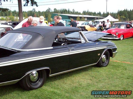 Black convertible with a white side stripe and chrome hubcaps parked on grass. Hood is open. Red car in background