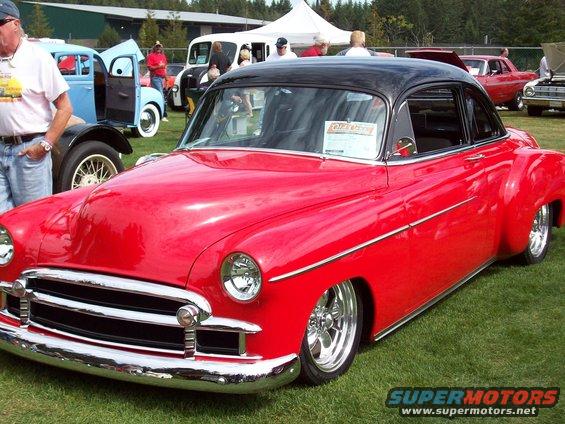 Red coupe with a black roof, lowered suspension, chrome grille, and custom wheels parked on grass