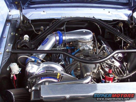 Engine bay of a blue classic car, likely a 1967 Ford Mustang, with a chrome intake, carburetor, and turbocharger