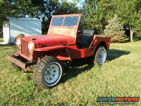 Red Willys Jeep with a rusty bumper and white rims, parked on grass