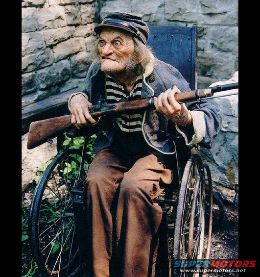 Old man with a rifle sits in a wheelchair next to a stone wall