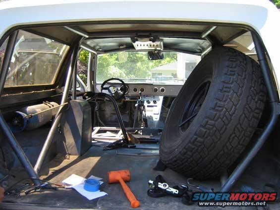 Inside a vehicle with a roll cage, spare tire, and tools on the floor. Orange mallet and blue tape are visible