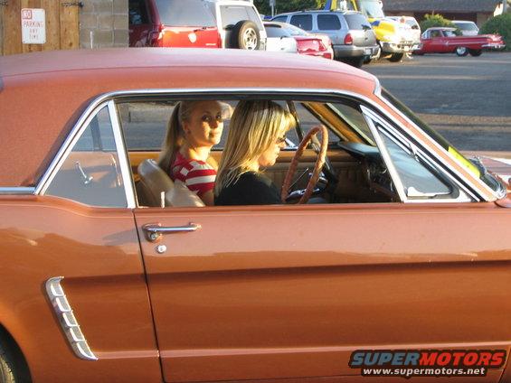 Orange 1965 Ford Mustang coupe with two blonde women inside, seen from the side