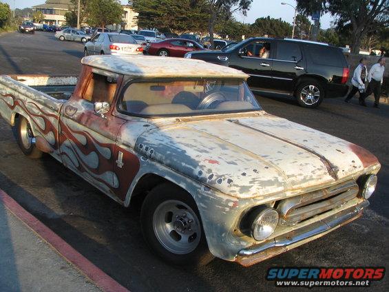 1957 Chevrolet pickup truck with custom flame paint and worn finish. Other vehicles and buildings in background