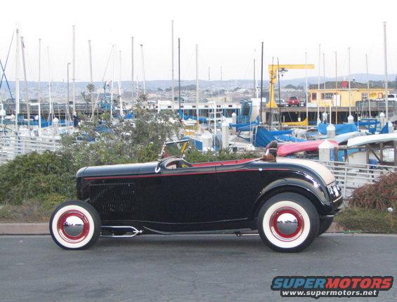 Black hot rod roadster with red trim and whitewall tires parked by a marina