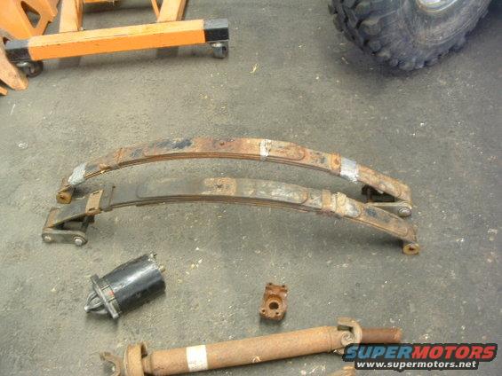 Leaf springs, starter motor, and driveshaft on concrete floor. Tire and orange jack stands in background