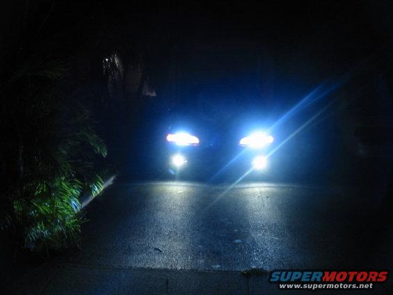 Two cars driving at night, their headlights are very bright. One car is on the left, the other on the right