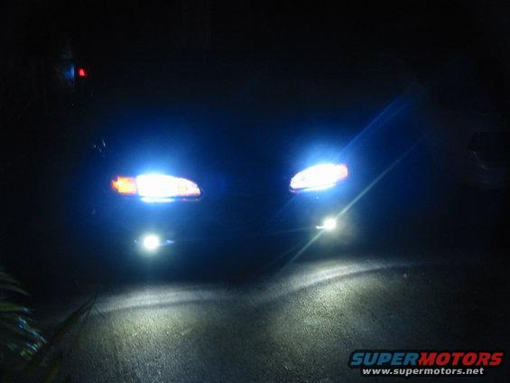 Two bright headlights illuminate a dark road at night. A small red light is visible in the upper left