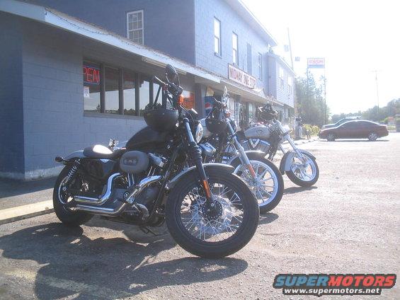 Three motorcycles parked in front of a blue building. The closest motorcycle is black with chrome accents. A red car is visible in the background