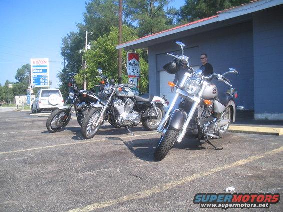 Silver motorcycle parked outside a building with other motorcycles and a gas station in the background