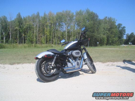 Harley-Davidson motorcycle parked on dirt road with trees in background. Watermark says SUPERMOTORS.NET
