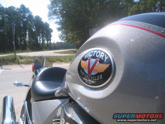 Close-up of Victory Motorcycles badge on a silver motorcycle. Road and trees in background